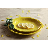 Textured Lemon Earthenware Plate in yellow, shaped like a whole lemon with a sculpted green leaf, dimpled peel-like surface, shown stacked with lemon slices on a neutral linen surface.