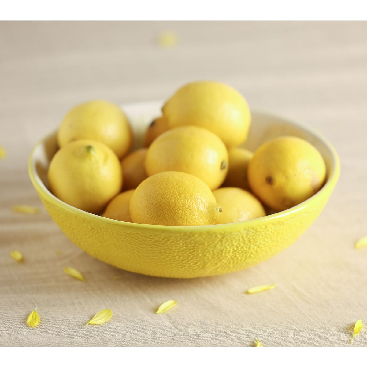 Round earthenware serving bowl with textured yellow peel-effect exterior, shown filled with whole lemons on a light fabric surface with scattered petals.