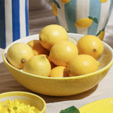 Round textured yellow earthenware serving bowl filled with whole lemons, styled on a table with striped jugs in the background.