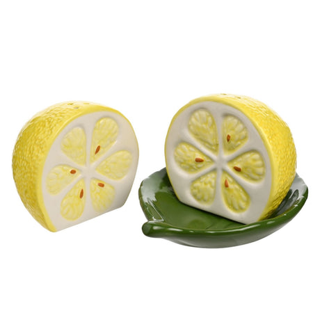 Lemon-shaped earthenware salt and pepper shaker set in yellow with white cut faces, resting on a green leaf-shaped ceramic dish, on a plain white background.