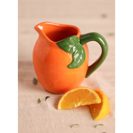 Small Citrus Fruit Earthenware Jug in Orange, shaped like an orange with dimpled texture, glossy glaze, green handle and leaf detail, shown with orange slices on a neutral surface.
