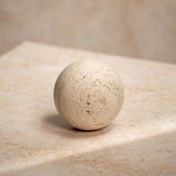 Travertine Sphere Ornament 9.7cm in brown, a round natural stone decorative accessory with pitted texture and tan veining, shown on a light travertine surface.