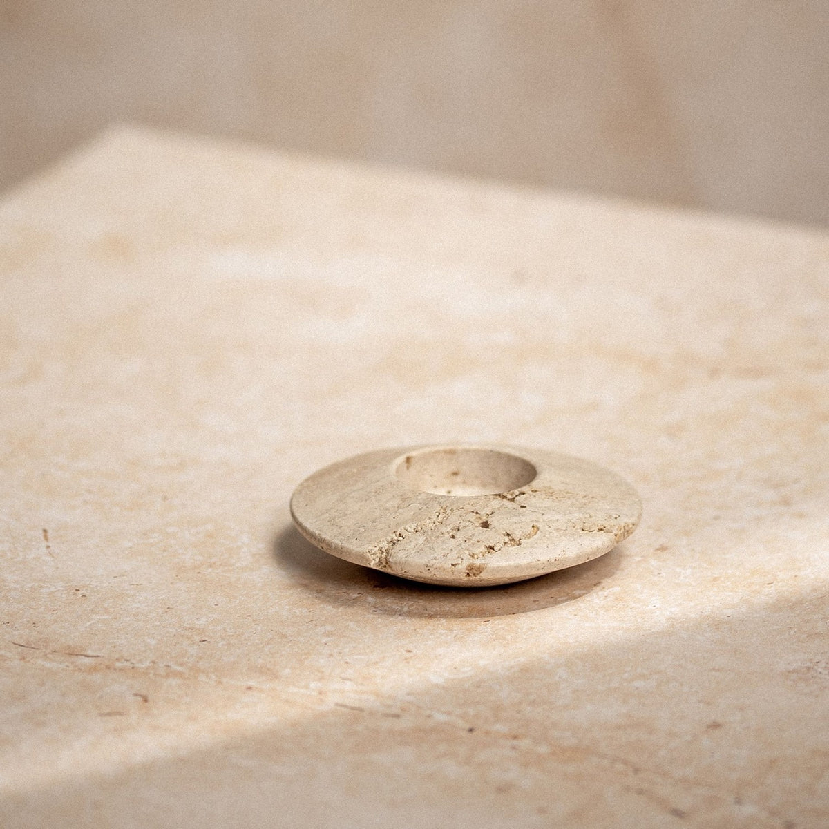 Travertine Disc Tea Light Candle Holder in brown, a round low stone disc with a central cup, natural pitted texture, shown on a light stone surface.