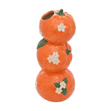 Ceramic vase shaped as three stacked oranges in glossy orange with dimpled peel texture, white citrus blossoms, green leaves and stalk, opening at top, on a plain white background.