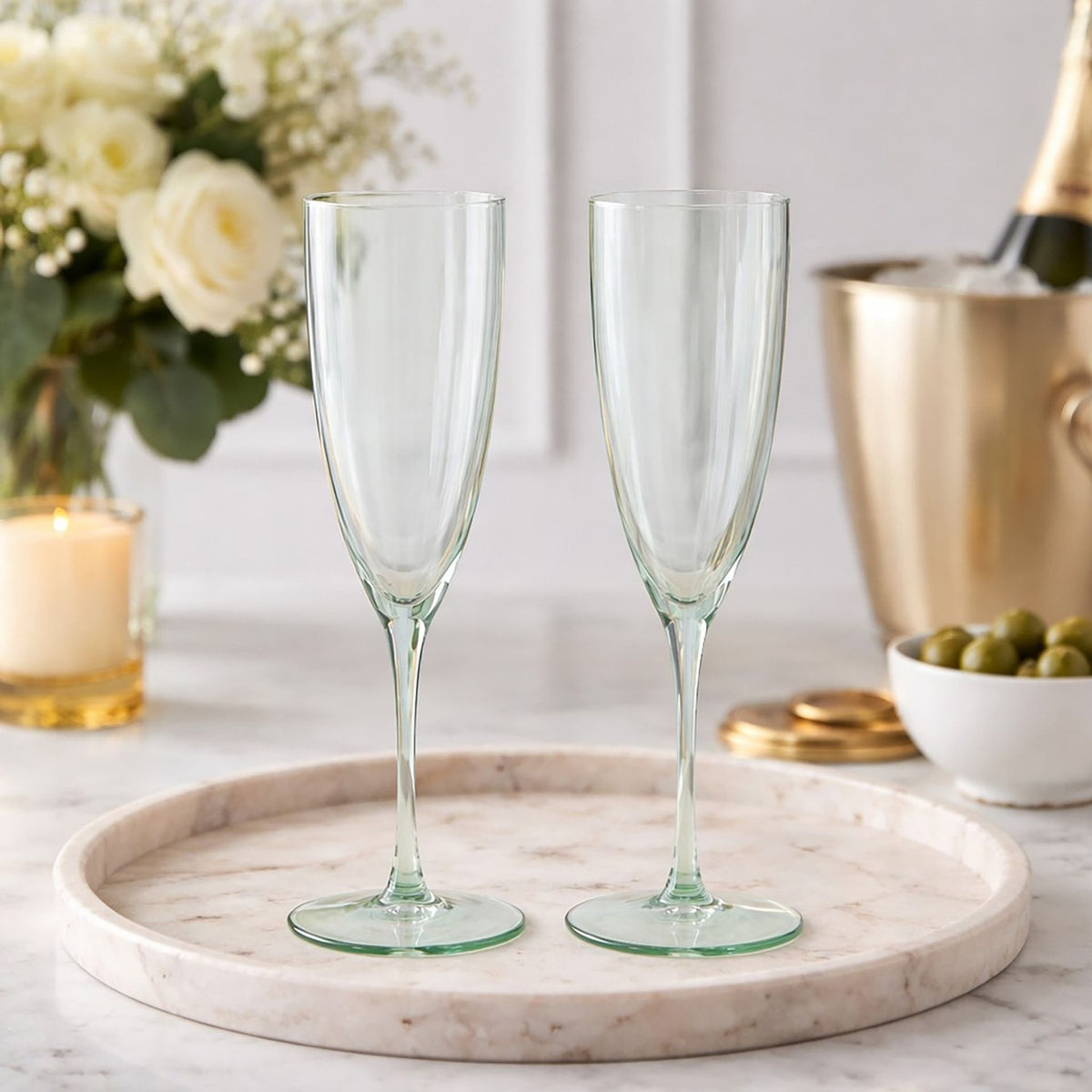 Set of 2 scalloped stemmed glass champagne flutes in pale green, shown on a marble tray with flowers and a champagne bucket in a dining setting.