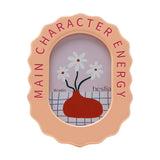 Wooden oval photo frame in pale orange with a wavy edge, "Main Character Energy" in pink capitals around the rim, holding a 6 x 4in daisy vase illustration on a white background.