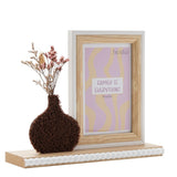 Wooden photo frame for a 6 x 4in portrait photo with natural wood surround and white edge, beside a textured dark brown vase of dried flowers, on a white background.