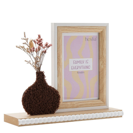 Wooden photo frame for a 6 x 4in portrait photo with natural wood surround and white edge, beside a textured dark brown vase of dried flowers, on a white background.