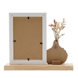 White 6x4in photo frame with wooden base, shown back-facing, beside a textured brown vase holding dried flowers, on a plain white background.