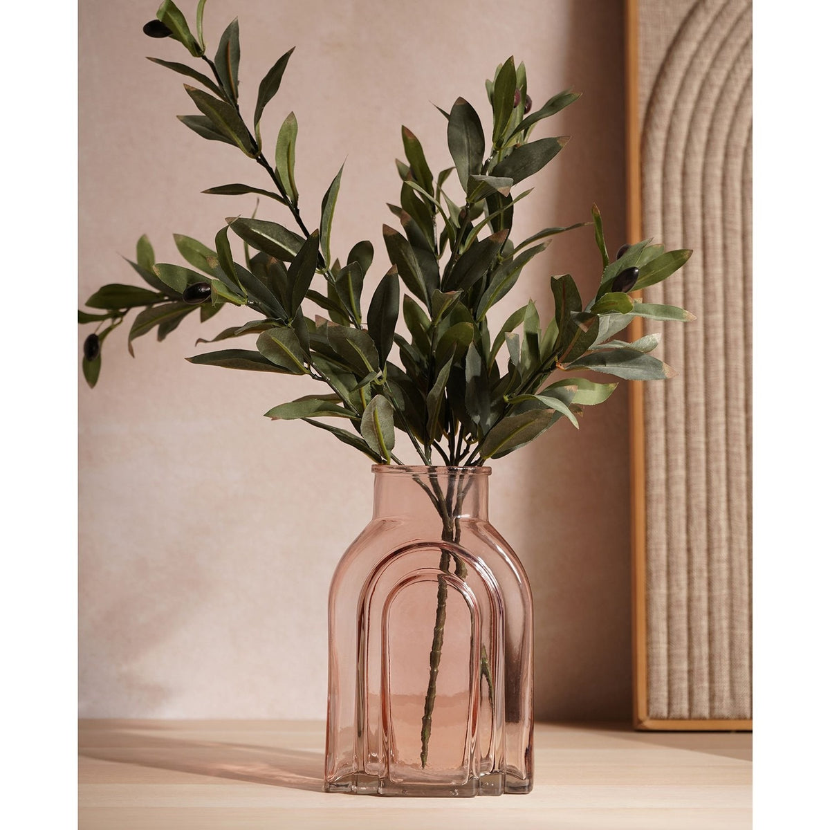Stepped arch glass vase in light pink with a narrow neck, holding green foliage stems on a wooden surface in a living room setting.