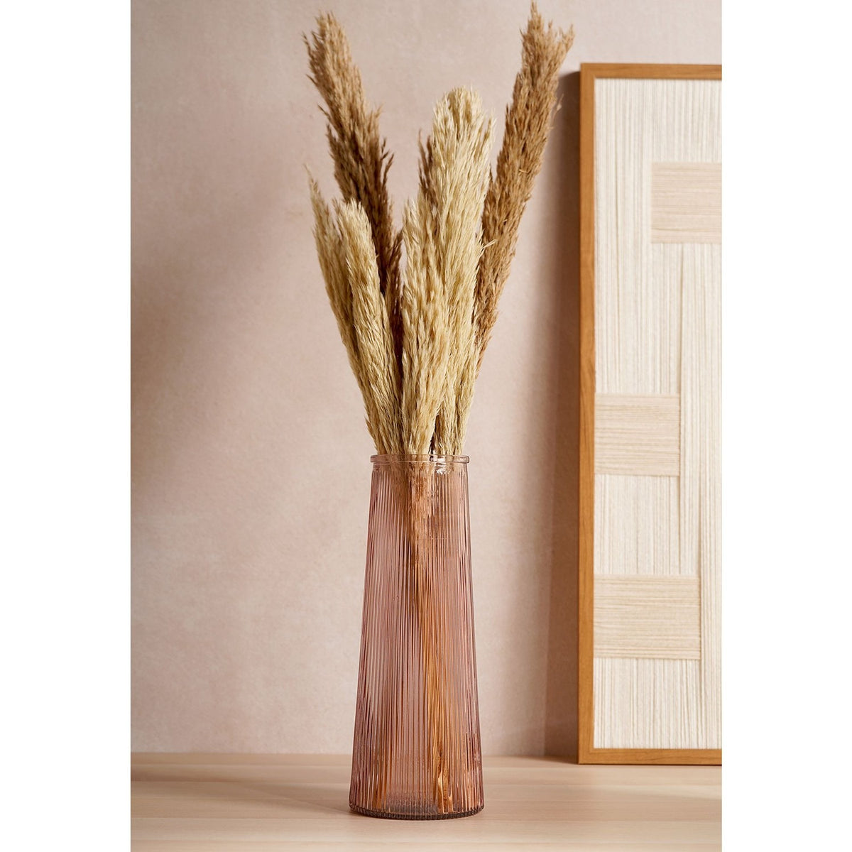 Tall ribbed tapered glass vase in pink, holding dried pampas grass stems on a wooden surface against a neutral wall and framed decor panel.
