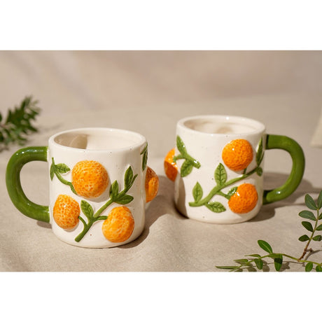 Two white earthenware mugs, 280ml, with raised dimpled orange fruits and green leafy branches, rounded green handles, shown on a neutral tablecloth setting.