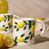 White ceramic mug with raised lemon and green leaf design, 600ml, shown beside fresh lemons on a neutral tabletop setting.