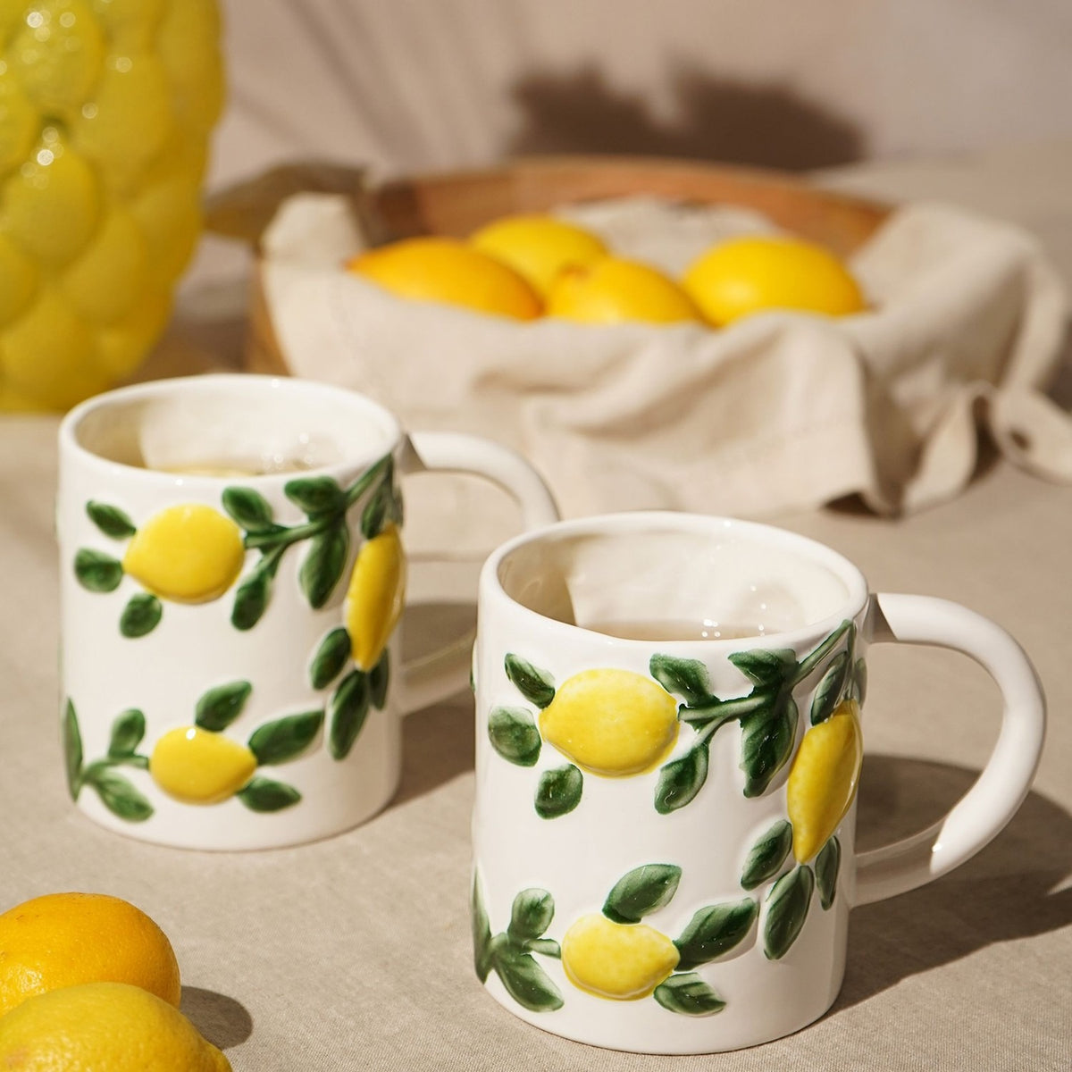 Two white ceramic mugs with raised yellow lemons and green leaf sprigs, filled with tea and shown beside fresh lemons on a linen-covered table.