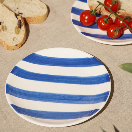 Round earthenware side plate, 16cm, with broad blue brushstroke stripes on white, glossy ceramic finish, styled on a linen tablecloth with bread and cherry tomatoes.