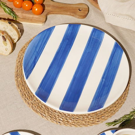 Round striped earthenware plate, 27.3cm, with bold cobalt blue and white stripes, shown on a woven placemat beside bread and cherry tomatoes.