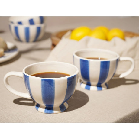 Round Striped Footed Earthenware Mugs 320ml in blue and white, filled with coffee on a dining table beside lemons and a matching bowl.