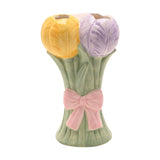 Ceramic vase shaped as a tulip bunch with yellow and lilac blooms on sage green stems tied with a raised pink bow, shown on a plain white background.