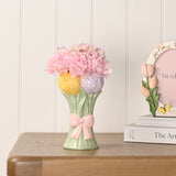 Ceramic vase shaped like a tulip bunch with yellow and lilac blooms, sage green stems, and a raised pink bow, styled with pink hydrangea stems on a wooden table.
