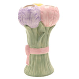 Ceramic vase shaped as a tulip bunch with yellow, lilac and pink blooms on sage green stems, tied with a raised pink bow, on a plain white background.
