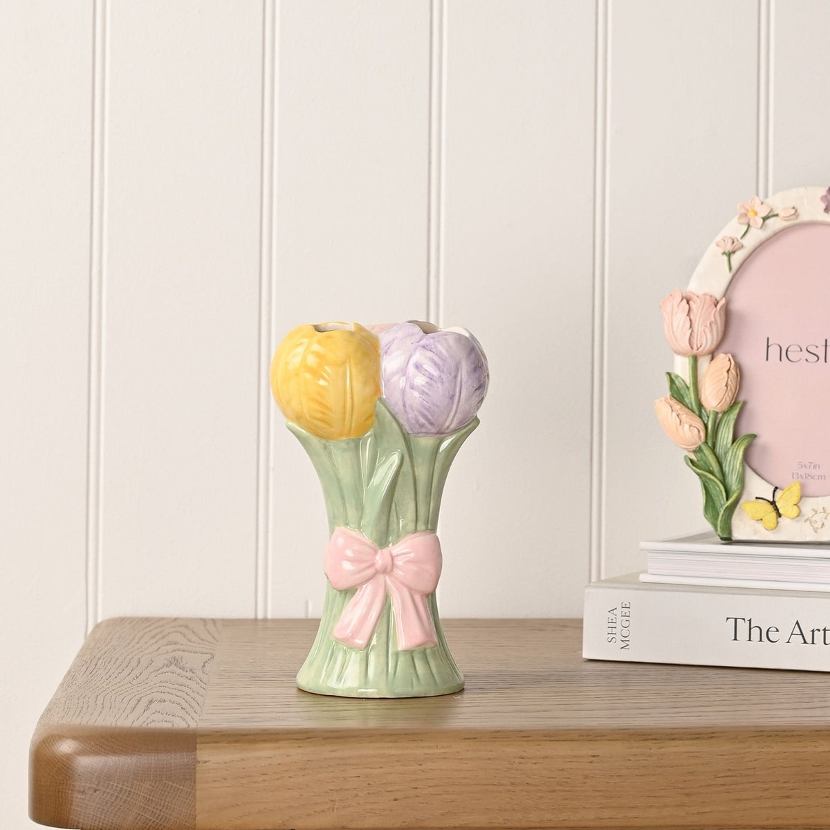 Ceramic vase shaped as a tulip bunch with yellow and lilac blooms on sage green stems tied with a pink bow, displayed on a wooden table beside books and a photo frame.