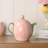 Blush Tulip earthenware teapot with pink petal-shaped body, sage green handle, ribbed spout, and bud knob lid, displayed on a wooden shelf.