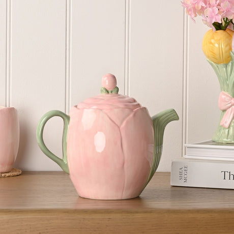 Blush Tulip earthenware teapot with pink petal-shaped body, sage green handle, ribbed spout, and bud knob lid, displayed on a wooden shelf.