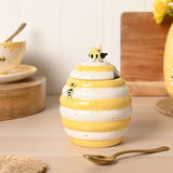 Beehive-shaped earthenware honey pot storage jar in ribbed white and yellow stripes, with a bee-shaped lid knob and a sculpted bee on the side, shown on a kitchen countertop.