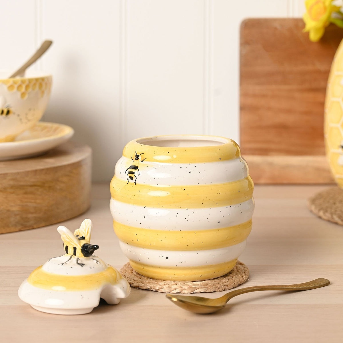 Bee Hive earthenware honey pot storage jar in ribbed white and yellow stripes with sculpted bee details, lid removed and placed beside it on a wooden kitchen surface.