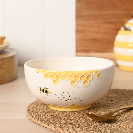 Cream round earthenware bowl with speckled texture, yellow honeycomb band along the rim and a raised bee with dotted flight path, shown on a woven mat beside a gold spoon.