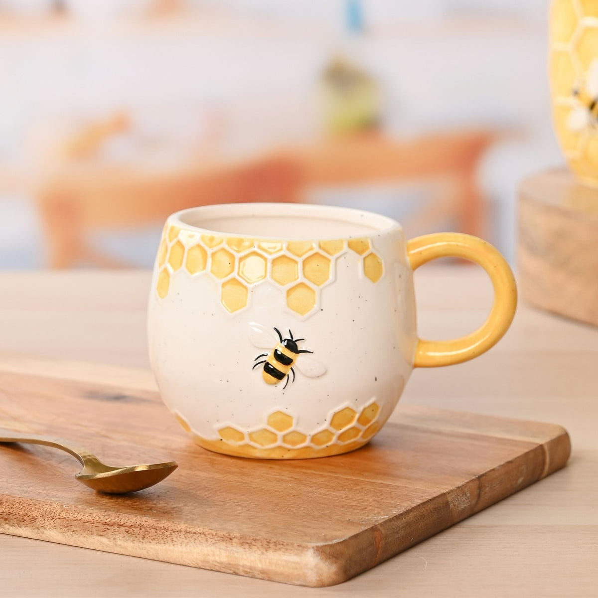 Earthenware mug in speckled cream with raised yellow honeycomb bands and a relief bee design, yellow loop handle, shown on a wooden board with a brass spoon.