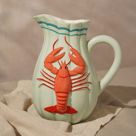 Hand-painted ceramic jug vase in pale green with vertical flutes, teal bands below the rim, and a raised coral red lobster in relief, shown on neutral linen fabric.