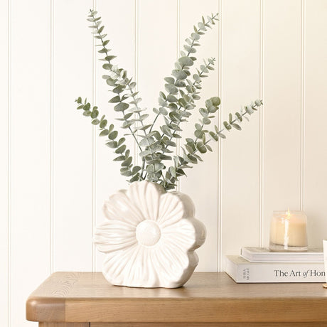 White glazed stoneware vase shaped like an open flower with textured centre, holding eucalyptus stems on a wooden table beside books and a candle.