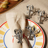 Set of 4 silver metal lobster napkin rings with twisted bands, detailed claws and tails, shown on a table with one fitted around a beige napkin on a patterned plate.