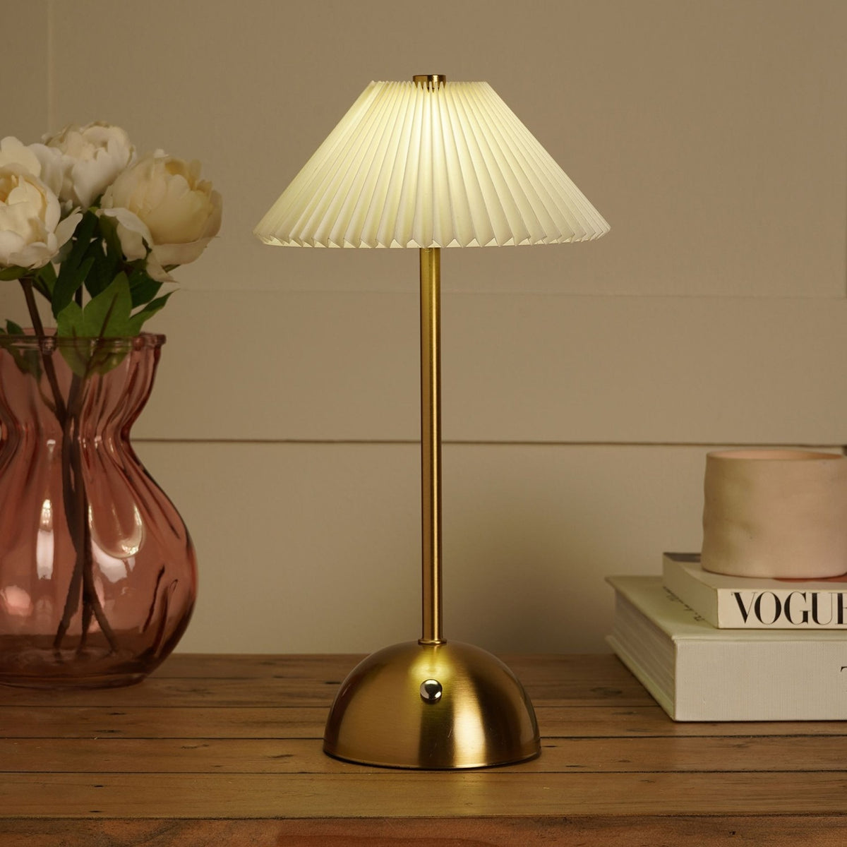 Pleated Shade USB LED Touch Table Lamp in bronze and cream, with a slim brushed bronze stem and dome base, shown lit on a wooden table beside books and a vase of flowers.