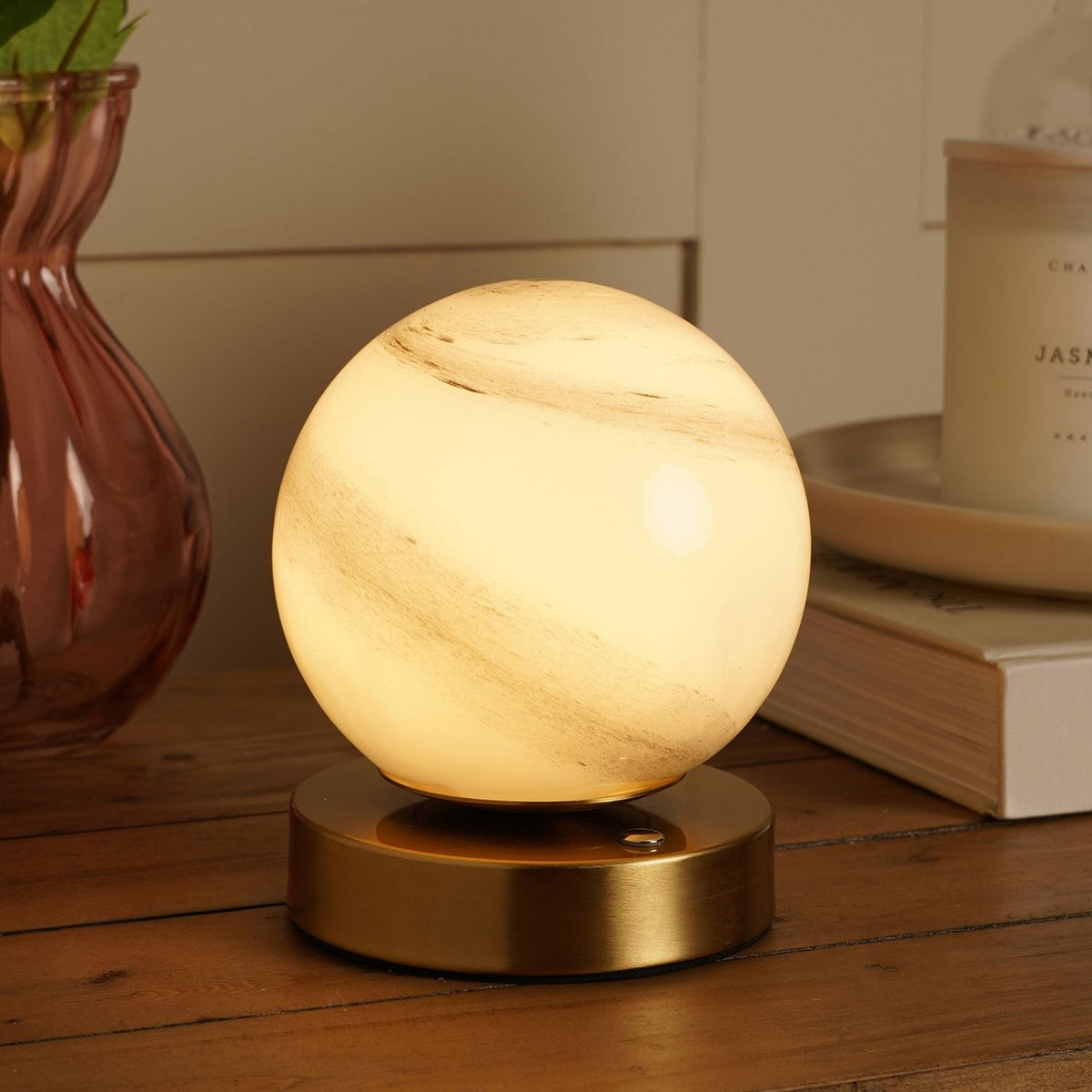 Marbled white glass ball table lamp glowing warm cream, with a round bronze-toned base, shown lit on a wooden surface beside a vase and candle.