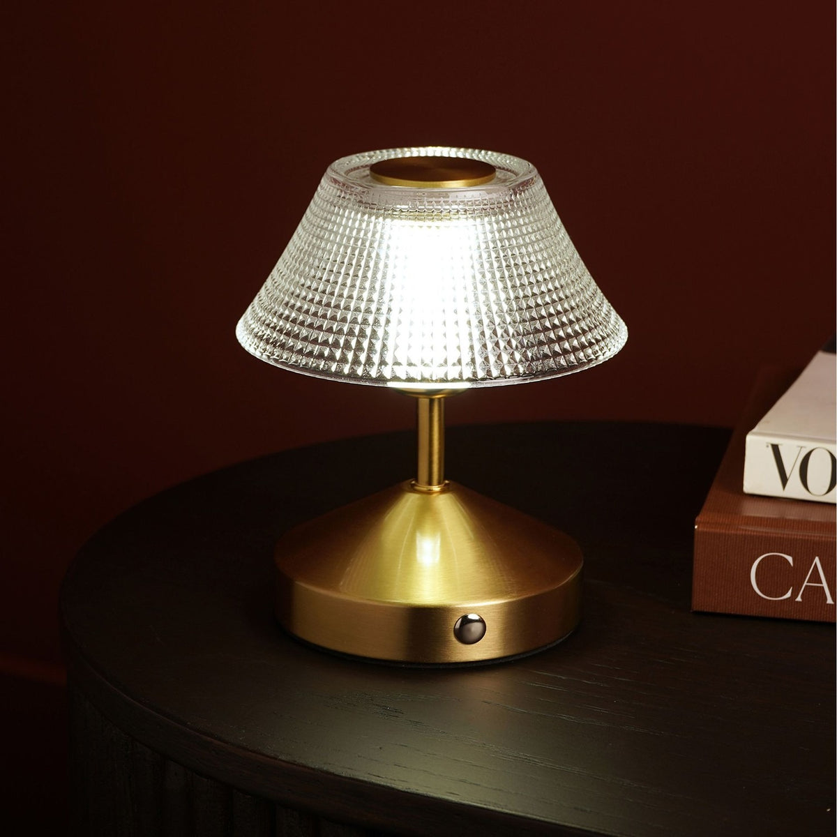 Small table lamp with a diamond-etched glass shade and brushed bronze base with touch control, shown lit on a dark table beside stacked books.