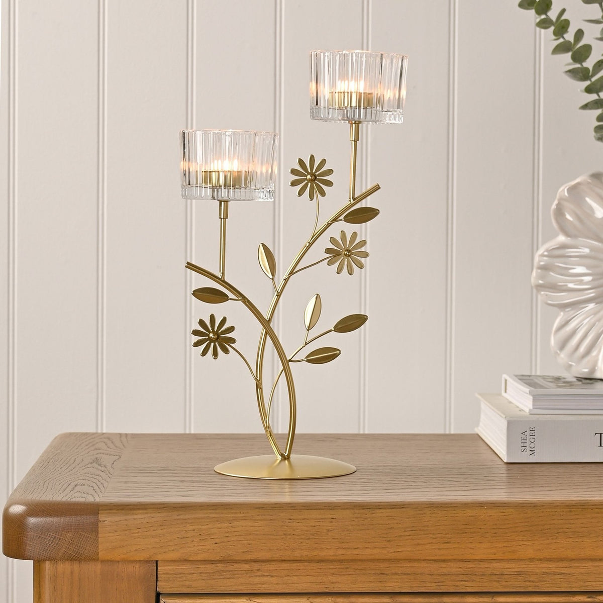 Floral Branch Double Metal Candle Holder in gold with clear ribbed glass cups, styled on a wooden sideboard with books and a decorative flower ornament.