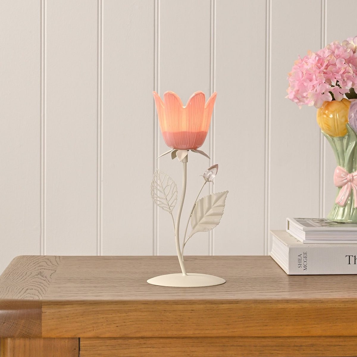 Floral Single Candle Holder with Metal Leaves in pink and cream, featuring a tulip-shaped cup on a slender stem with leaf details, shown lit on a wooden sideboard beside books and a flower vase.