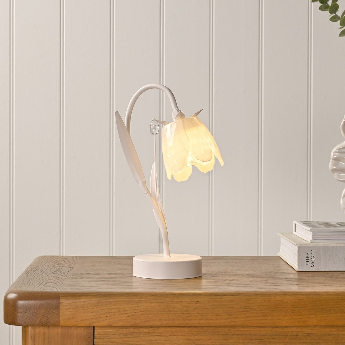 Floral Bell Shade Battery LED Table Lamp in cream, with curving stem, leaf details, and round base, shown lit on a wooden table against a white panelled wall.