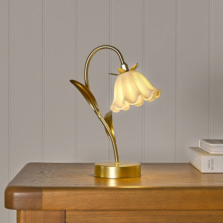 Floral Stem LED Table Lamp with gold curved stem, leaf accents, round base, and a fluted bell-shaped shade, shown lit on a wooden sideboard.