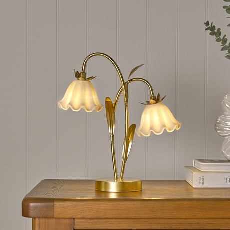 Floral 2 Stem LED Table Lamp with bell-shaped pale pink glass shades atop gold curving stems and leaf details, on a round gold base, shown lit on a wooden sideboard.