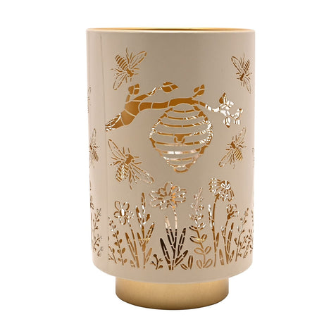 Cream metal LED table lamp with cut-out bee, beehive and wildflower design, glowing gold interior and matching gold base, shown on a plain white background.