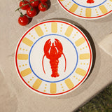Round cream dinner plate with a red lobster at the centre, blue ring border, yellow dash-and-dot pattern, and a red rim edge, shown on a table beside cherry tomatoes.
