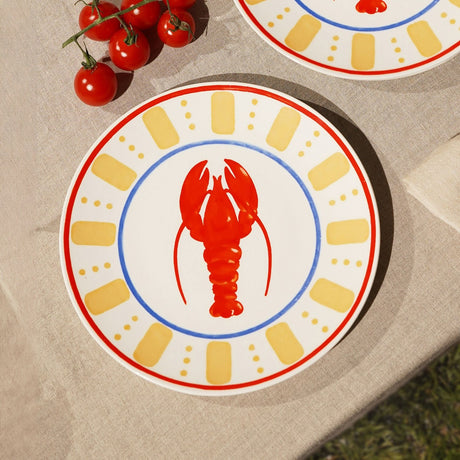 Round cream dinner plate with a red lobster at the centre, blue ring border, yellow dash-and-dot pattern, and a red rim edge, shown on a table beside cherry tomatoes.
