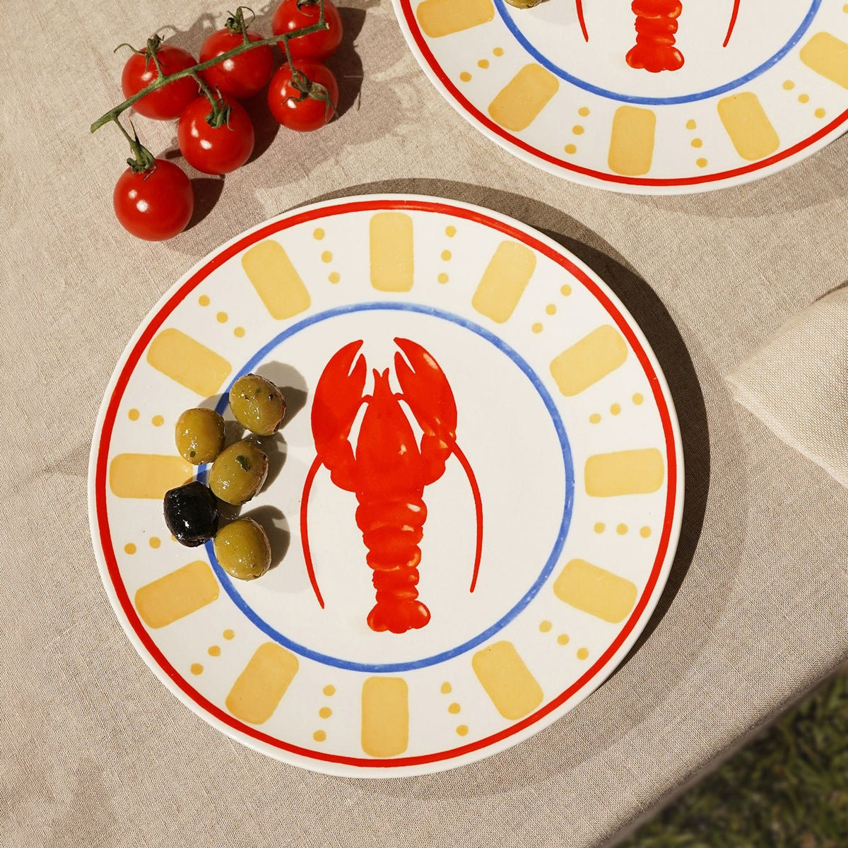 Round cream dinner plate with a red lobster design, blue ring border, yellow dash and dot pattern, and red rim, shown outdoors with olives and cherry tomatoes.