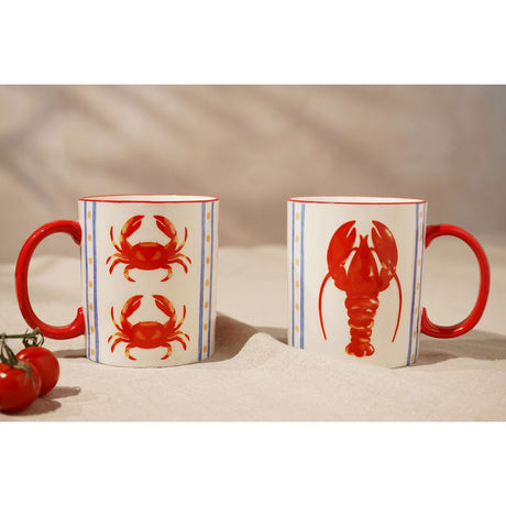 Set of 2 crab and lobster mugs in cream with red handles and rims, featuring red crab and lobster illustrations between blue stripes, shown with fresh tomatoes on a neutral tabletop.
