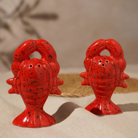 Speckled Lobster Salt & Pepper Set in Red, a pair of ceramic pots shaped as upright lobsters with raised claws forming a loop, glossy red finish with dark speckles, shown on a neutral woven surface.