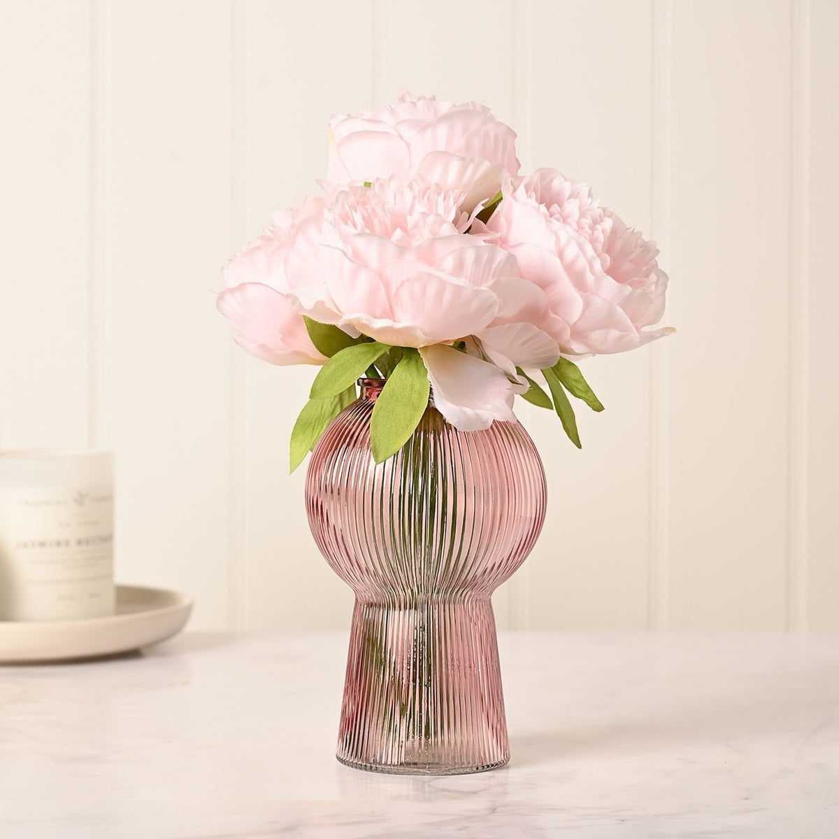 Pink ribbed glass vase with round balloon body and flared pedestal base, holding pale pink peonies, styled on a marble surface.