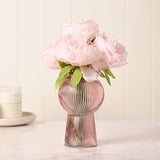 Pink ribbed glass vase with round balloon body and flared pedestal base, holding pale pink peonies, styled on a marble surface.